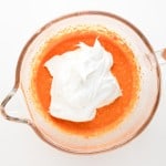 A glass mixing bowl with orange batter and a dollop of whipped egg whites on top, viewed from above on a white background.