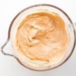 A glass mixing bowl filled with a creamy, light orange batter or mixture, viewed from above on a white surface.