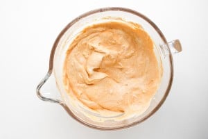 A glass mixing bowl filled with a creamy, light orange batter or mixture, viewed from above on a white surface.