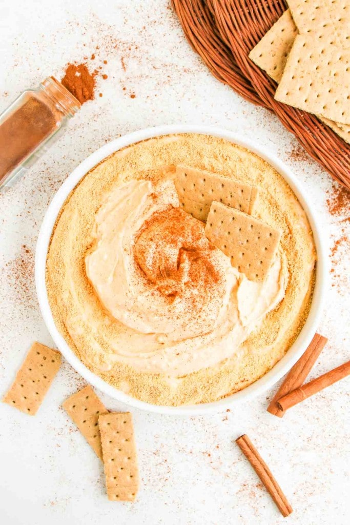 A bowl of creamy dip topped with graham cracker crumbs and cinnamon, surrounded by graham crackers and cinnamon sticks on a white surface.