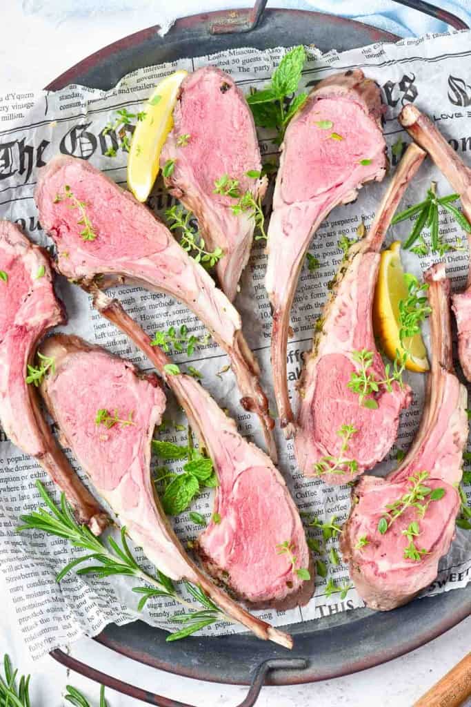 Sliced Rack of Lamb, cooked medium-rare, arranged on newspaper with fresh herbs and lemon wedges for garnish.