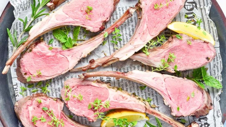 Several slices of medium-rare rack of lamb, garnished with fresh herbs and lemon wedges, are artfully arranged on newspaper print paper.