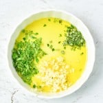 A white bowl filled with olive oil, chopped fresh herbs, and minced garlic&mdash;perfect for drizzling over a succulent rack of lamb&mdash;rests on a light marble surface.