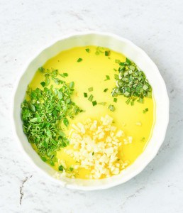 A white bowl filled with olive oil, chopped fresh herbs, and minced garlic—perfect for drizzling over a succulent rack of lamb—rests on a light marble surface.