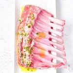 A raw rack of lamb drizzled with oil and topped with chopped garlic and herbs sits on a white rectangular plate.