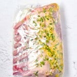 Raw rack of lamb marinated with herbs and oil, sealed in a clear plastic bag and ready for cooking, sits on a light-colored surface.