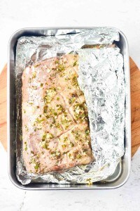 A seasoned, scored pork roast sits in a foil-lined baking pan, partially covered with foil, on a wooden surface—much like how you might prepare a rack of lamb for a savory feast.