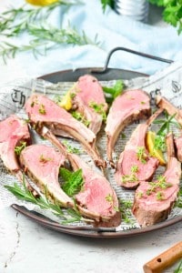 A round tray lined with newspaper holds six slices of succulent rack of lamb, garnished with fresh herbs and lemon wedges.