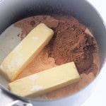 Two sticks of butter, cocoa powder, and liquid cream in a saucepan, ready to be mixed and heated.