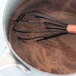 A whisk stirs a pot of chocolate mixture on a stove, showing swirls in the liquid.