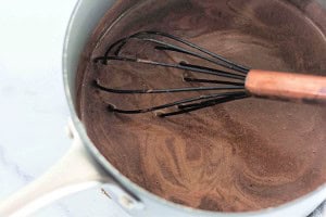 A whisk stirs a pot of chocolate mixture on a stove, showing swirls in the liquid.