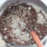 A saucepan containing a chocolate mixture and dry flour being mixed with a whisk on a white countertop.