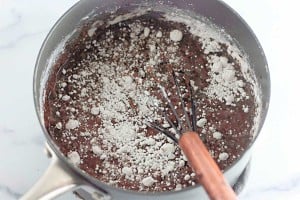 A saucepan containing a chocolate mixture and dry flour being mixed with a whisk on a white countertop.