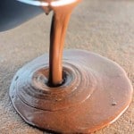 A stream of melted chocolate is being poured from a bowl onto a flat surface, creating a smooth, glossy pool.