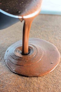 A stream of melted chocolate is being poured from a bowl onto a flat surface, creating a smooth, glossy pool.