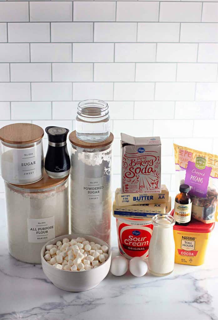 Baking ingredients on a counter, including flour, sugar, powdered sugar, baking soda, cocoa, sour cream, butter, eggs, white chocolate chips, vanilla extract, and pecans.