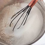 A metal mixing bowl containing white flour and granulated sugar being whisked together with a wire whisk.