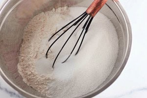 A metal mixing bowl containing white flour and granulated sugar being whisked together with a wire whisk.