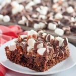 A square piece of chocolate cake topped with marshmallows, pecans, and chocolate drizzle on a white plate, with more cake in the background.