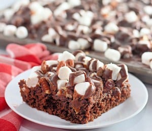 A square piece of chocolate cake topped with marshmallows, pecans, and chocolate drizzle on a white plate, with more cake in the background.
