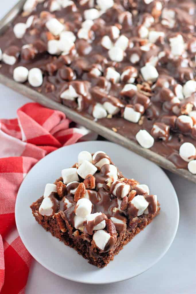 A brownie topped with marshmallows, pecans, and chocolate frosting sits on a white plate, with a pan of similar brownies in the background.