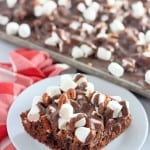 A chocolate dessert bar topped with mini marshmallows and nuts on a white plate, with a tray of similar bars and a red checkered cloth in the background.