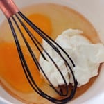 A metal whisk in a white bowl with two raw eggs, a dollop of sour cream, and some liquid ingredients.