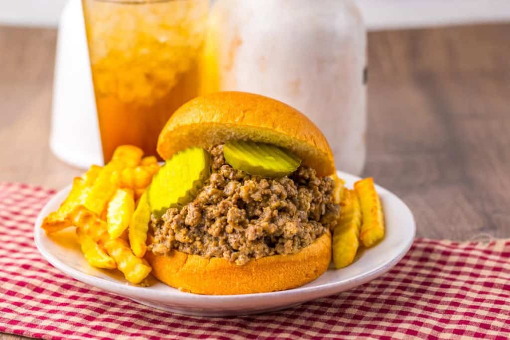 A sandwich with loose ground beef and pickles on a bun, served with crinkle-cut fries on a white plate, with a drink in the background.