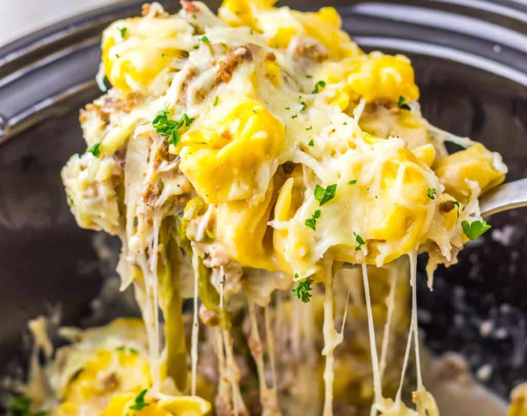 A close-up of cheesy tortellini pasta being lifted with a spoon, topped with melted cheese and sprinkled with chopped herbs&mdash;perfect for fans of Slow Cooker recipes.
