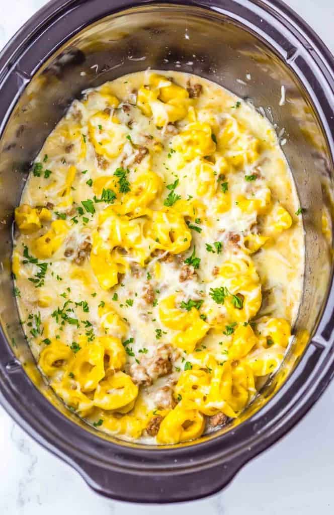 Cheese tortellini, sausage, and creamy sauce garnished with chopped parsley in a slow cooker.