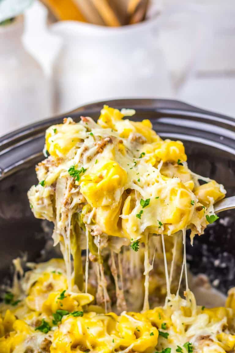 A spoon lifts cheesy tortellini pasta with herbs from a slow cooker, showing melted cheese stretching between the pasta pieces.