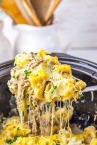 A spoon lifts a serving of cheesy tortellini with herbs from a slow cooker, showing melted cheese and pasta.