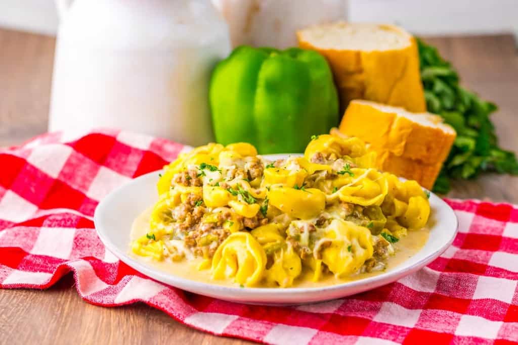 A plate of tortellini pasta with a creamy meat sauce sits on a red checkered cloth, with a green bell pepper, parsley, and sliced bread in the background.