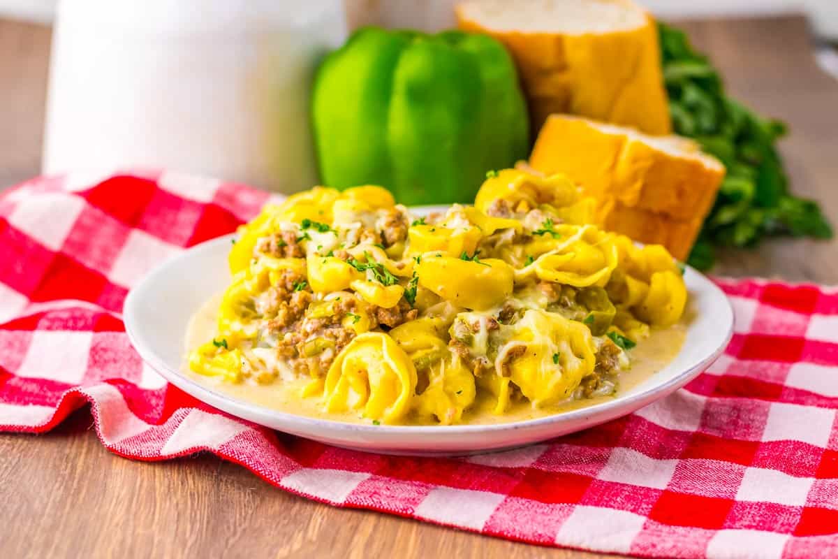 A plate of cheesy tortellini with ground meat served on a red checkered cloth, with bread, a green bell pepper, and greens in the background.
