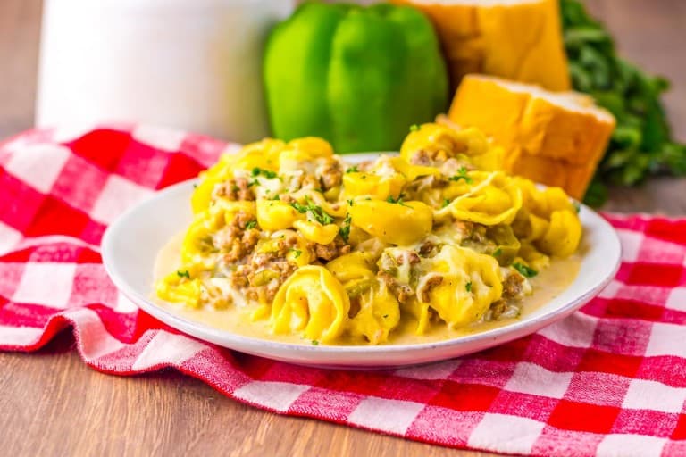 A plate of tortellini pasta with a creamy meat sauce sits on a red and white checkered cloth, with bread and a green bell pepper in the background.