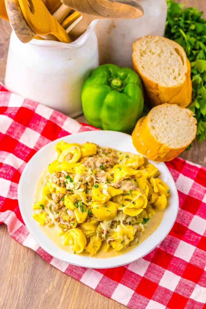 A plate of cheesy tortellini with ground meat served on a red checkered cloth, with bread, a green bell pepper, and greens in the background.
