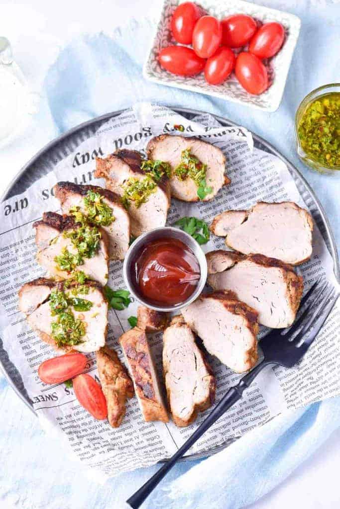 Sliced grilled pork tenderloin garnished with herbs, served with barbecue sauce, grape tomatoes, and green relish on a round plate lined with newspaper.