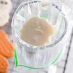 A plastic piping bag placed in a glass is filled with white icing, ready for decorating Halloween cookies. Decorated cookies and a cooling rack are visible in the background.