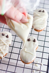 A hand pipes white icing onto a Halloween cookie shaped like a mummy, with chocolate chips for eyes, resting on a cooling rack.