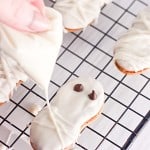 A hand pipes white icing onto a Halloween cookie shaped like a mummy, with chocolate chip eyes, resting on a cooling rack alongside other festively decorated Halloween cookies.