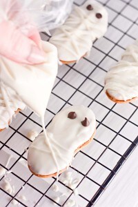 A hand pipes white icing onto a Halloween cookie shaped like a mummy, with chocolate chip eyes, resting on a cooling rack alongside other festively decorated Halloween cookies.