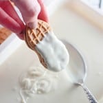 A hand dips a peanut-shaped Halloween cookie halfway into melted white chocolate in a shallow dish with a spoon.