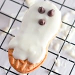 A peanut-shaped Halloween cookie covered in white icing with three chocolate chips on top, resembling a ghost, sits on a cooling rack.