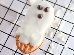 A peanut-shaped Halloween cookie covered in white icing with three chocolate chips on top, resembling a ghost, sits on a cooling rack.
