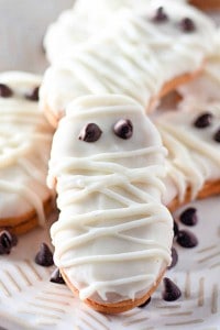 A Halloween cookie decorated to look like a mummy, covered in white icing with chocolate chip eyes, is placed on a plate surrounded by other spooky Halloween cookies.