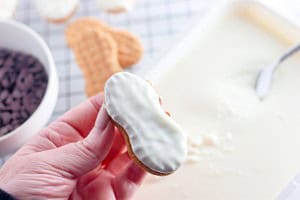 A hand holds a peanut-shaped cookie coated with white icing; chocolate chips, more Halloween cookies, and a tray of icing are visible in the background.
