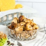 A glass bowl filled with grilled chicken pieces sits on a striped cloth, with tortilla chips and a plate of rice, beans, and lettuce in the background.
