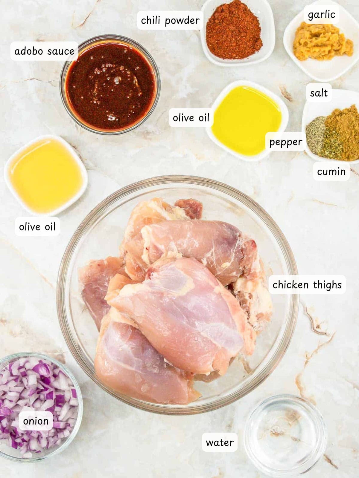 A bowl of raw chicken thighs surrounded by labeled ingredients&mdash;olive oil, adobo sauce, chili powder, garlic, salt, pepper, cumin, onion, and water&mdash;sit on a marble surface, ready to make flavorful copycat Chipotle chicken.