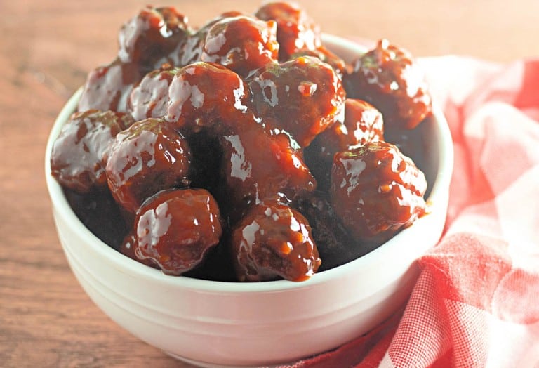 A white bowl filled with Slow Cooker Hot Honey Meatballs, coated in a thick, shiny barbecue sauce, sits on a wooden surface next to a red and white cloth.
