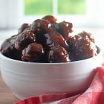 A white bowl filled with Slow Cooker Hot Honey Meatballs sits on a wooden surface next to a red and white checkered cloth, with a blurred window in the background.